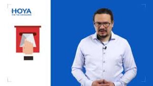 Mężczyzna w okularach prezentujący materiały o wadach wzroku dzieci, logo Hoya w tle, niebieska ściana.