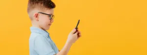 Chłopiec w okularach patrzy na smartfon na tle żółtej ściany, kontekst: krótkowzroczność u dzieci.