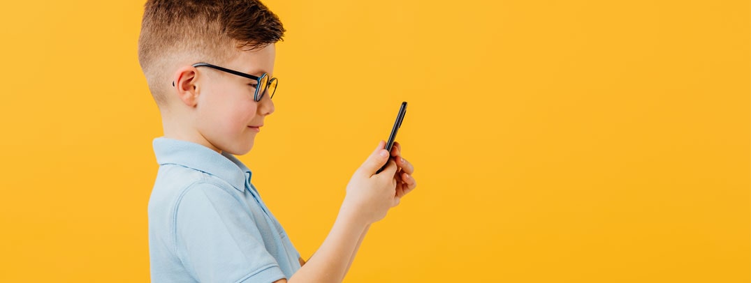 Chłopiec w okularach patrzy na smartfon na tle żółtej ściany, kontekst: krótkowzroczność u dzieci.