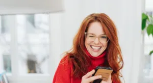 Kobieta w czerwonym swetrze z okularami, uśmiecha się, trzymając smartfon, w tle jasne wnętrze.