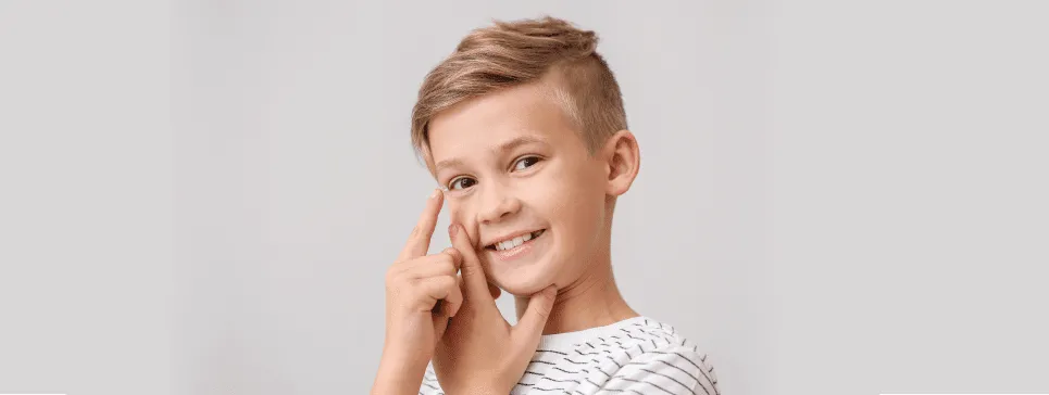 Chłopiec z uśmiechem w białej koszulce wskazuje na swoje oko, promując świadomość wad wzroku u dzieci.