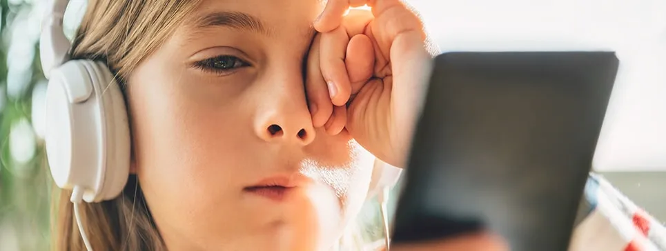 Dziecko w słuchawkach patrzy na smartfon, trzymając dłoń przy oku, nawiązuje do krótkowzroczności.